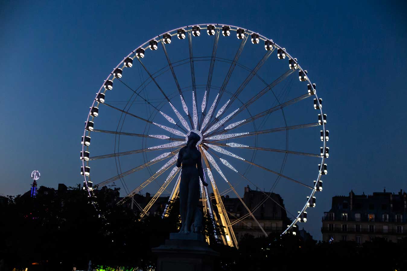 Imagen de una feria al caer la noche. La presencia de una estatua a contraluz, contrasta con la estampa de la noria iluminada al fondo
